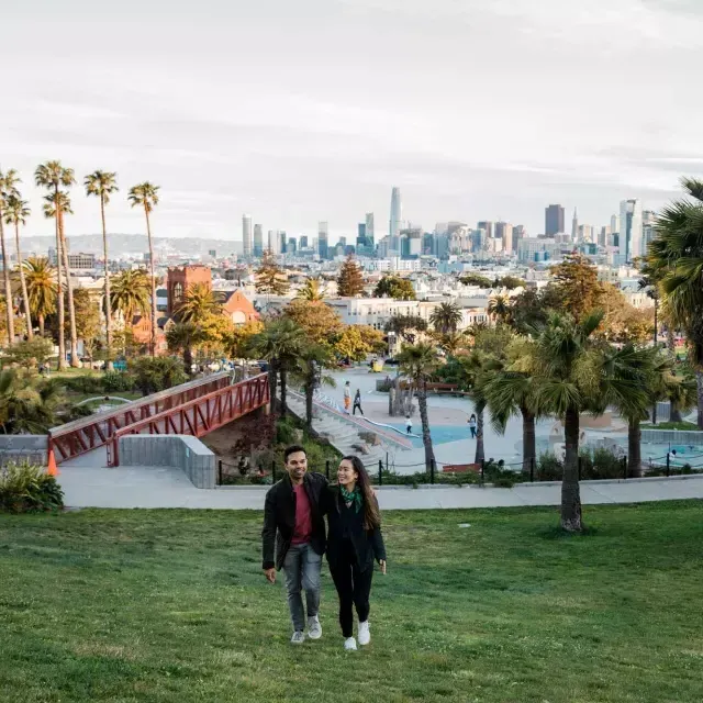 Una coppia cammina verso la telecamera con Dolores Park e lo skyline di 威尼斯人官网平台app dietro di loro.